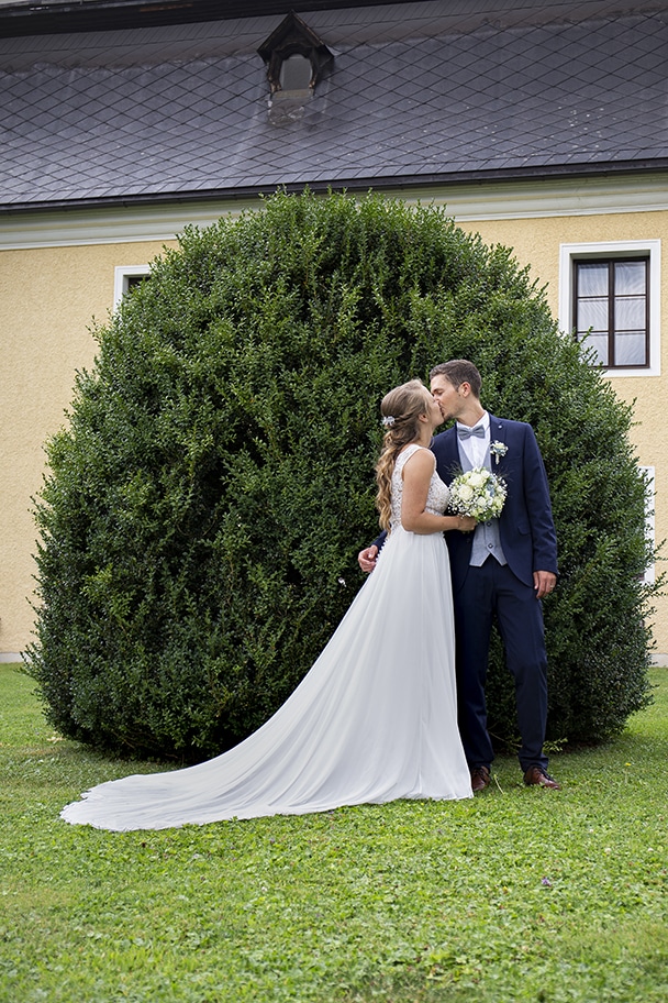 Braut und Bräutigam stehen vor einer großen, üppigen Hecke und küssen sich. Die Braut trägt ein fließendes weißes Kleid und der Bräutigam einen dunklen Anzug, was ihr Hochzeitsglück perfekt einfängt. Sie stehen auf einer Wiese mit einem beigefarbenen Gebäude im Hintergrund.