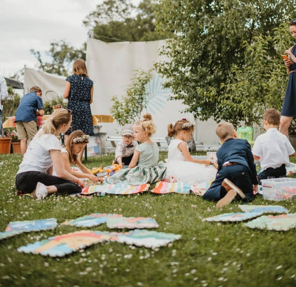 Kinder und Erwachsene versammeln sich draußen auf einer Rasenfläche und sitzen auf bunten Matten. Einige Kinder spielen mit Spielzeug, während andere sich mit Aktivitäten beschäftigen, die den Festtagen noch mehr Freude verleihen. Bäume und Grünflächen schaffen die Kulisse für diesen lebhaften Anlass, der sich perfekt für jede Hochzeitsfeier eignet.