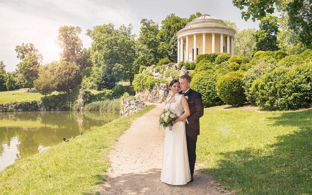 Braut und Bräutigam stehen auf einem Weg an einem ruhigen See, umgeben von üppigem Grün. Als Teil ihrer sorgfältigen Hochzeitsplanung ziert ein runder Pavillon mit Säulen den Hintergrund. Die Braut umarmt sich zärtlich und hält einen Strauß weißer Blumen unter dem klaren, sonnigen Himmel.