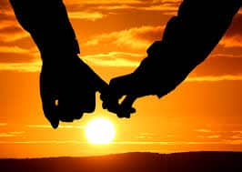 Die Silhouetten der Hände, die sich sanft vor einem leuchtend orangefarbenen Abendhimmel halten, bilden die perfekte Kulisse für eine romantische Hochzeit, während die Sonne am Horizont untergeht.