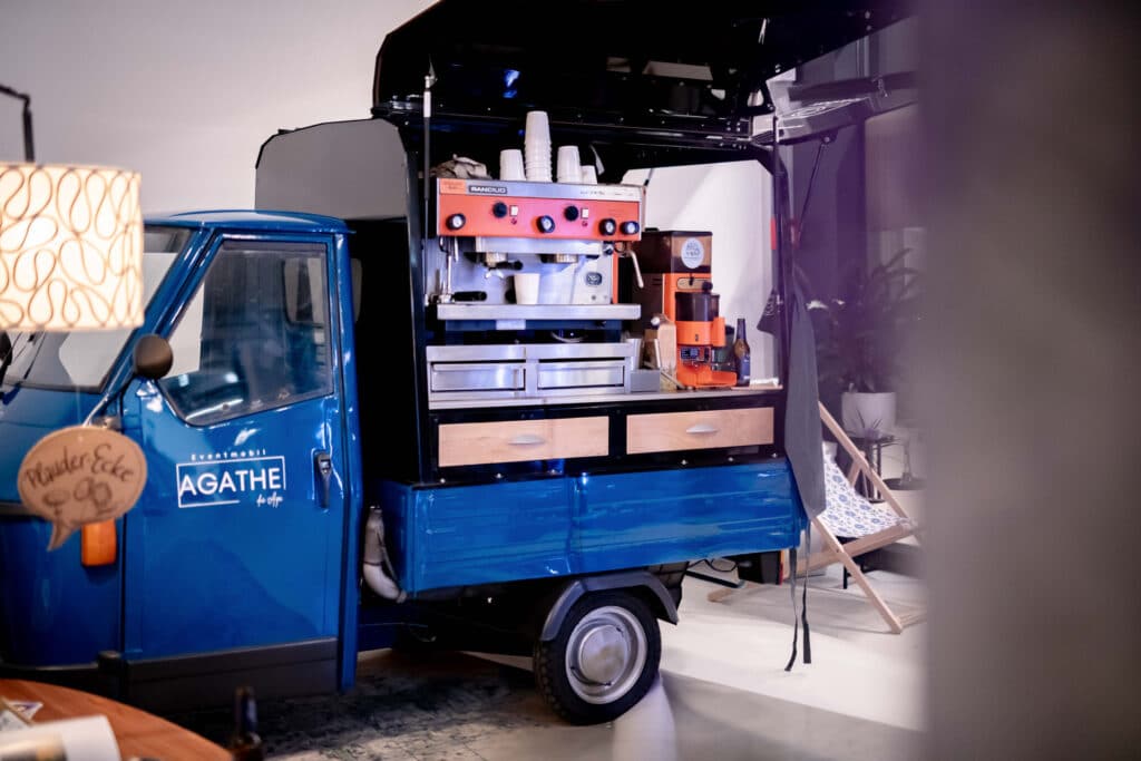 Ein kleiner blauer mobiler Kaffeewagen mit offener Klappe, ausgestattet mit einer orangefarbenen Espressomaschine, Tassen und Schubladen. Perfekt für Hochzeitsfeiern im Innenbereich. Stellen Sie sich dieses gemütliche Setup bei Ihrer Hochzeit vor, mit einer Hängematte und einer gemütlichen Lampe, die dem Hintergrund Charme verleiht.