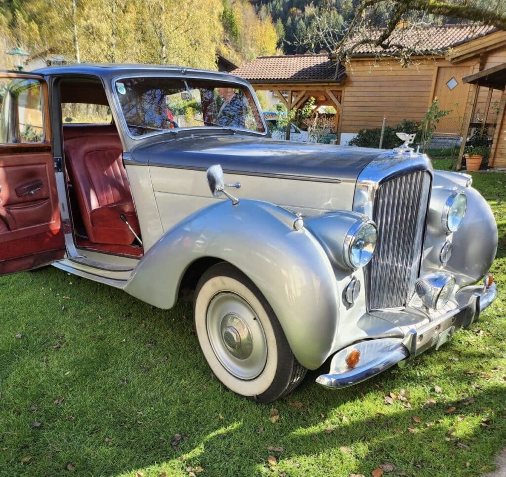 Ein silberner Oldtimer mit Weißwandreifen steht auf einer Wiese, perfekt für den Eingang einer Hochzeit. Die Fahrertür ist offen und gibt den Blick auf die roten Sitze im Inneren frei. Ein Holzhaus und Bäume stehen wunderschön im Hintergrund unter einem klaren Himmel und verleihen jeder Hochzeitsplanung Charme.