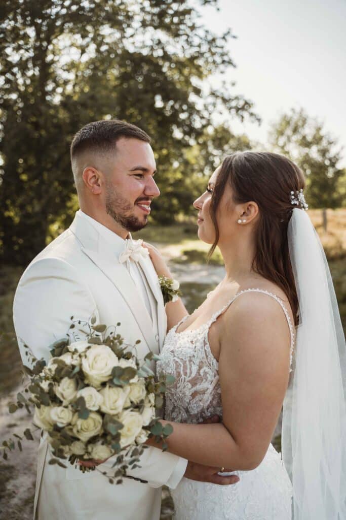 Bei ihrer Hochzeit stehen sich Braut und Bräutigam im Freien gegenüber. Die Braut hält einen Strauß weißer Rosen in der Hand und trägt ein Spitzenkleid mit Schleier, während der Bräutigam einen weißen Anzug mit Fliege trägt. In Sonnenlicht getauchte Bäume bieten die perfekte Kulisse für diesen zauberhaften Moment des Heiratens.
