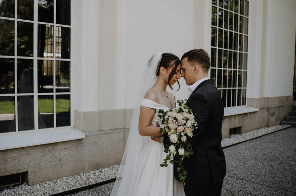 Braut und Bräutigam stehen dicht beieinander draußen vor einem weißen Gebäude mit hohen Fenstern. Die Braut hält einen Strauß rosafarbener und weißer Blumen und beide sind in formelle Hochzeitskleidung gekleidet. Sie teilen einen intimen Moment, der ihre sorgfältige Hochzeitsplanung widerspiegelt.