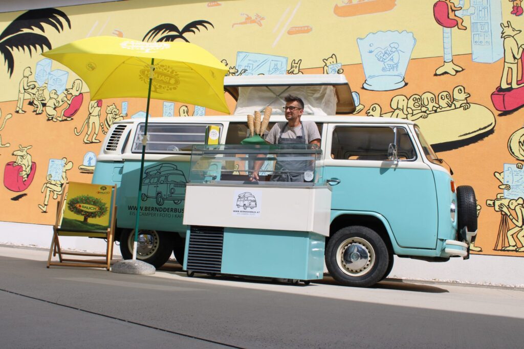 Ein mintgrüner und weißer Oldtimer-Van, perfekt für eine Hochzeit, steht vor einem farbenfrohen Wandgemälde. Der Van wurde in einen Eisstand mit einem lächelnden Verkäufer umgebaut. Ein Sonnenschirm spendet Schatten und daneben hängt ein Schild aus Kreidetafel. Ideal, um Ihrer Hochzeitsplanung Charme zu verleihen!