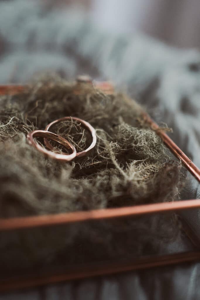 Zwei goldene Ringe ruhen auf einem Moosbett, präsentiert in einem quadratischen Kupferrahmen. Perfekt für alle, die sich intensiv mit Hochzeitsplanung beschäftigen. Das Bild verfügt über einen unscharfen Hintergrund, der die Ringe und das Moos hervorhebt und die Essenz des Heiratszaubers einfängt.