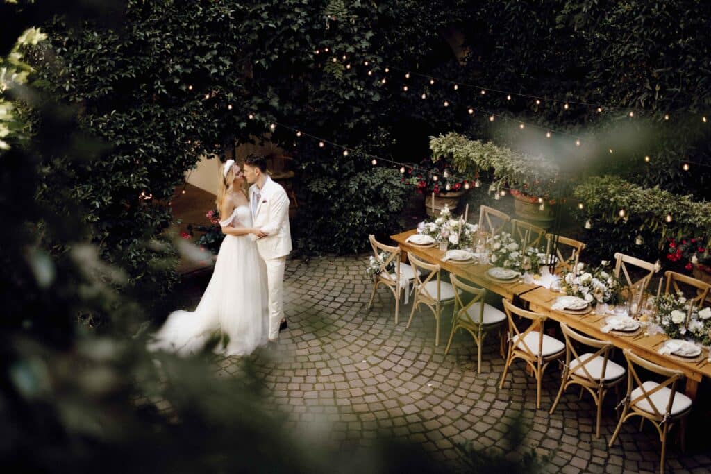 Ein Paar in Hochzeitskleidung umarmt sich auf einem Kopfsteinpflasterweg in einer Gartenlandschaft, ihr liebevoller Moment wird vom Schein der Lichterketten und üppigem Grün eingerahmt. In der Nähe erwartet sie ein wunderschön dekorierter Tisch, der die Eleganz der sorgfältigen Hochzeitsplanung für ihre intime Hochzeitsfeier widerspiegelt.