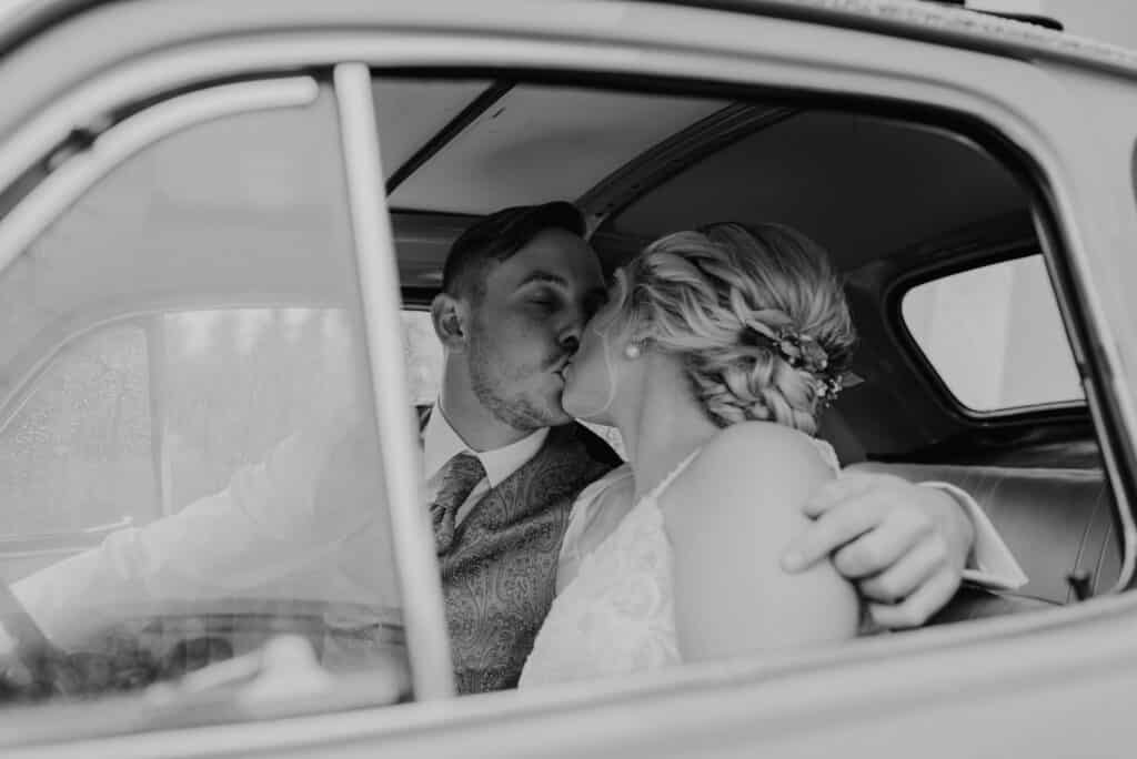 Ein Schwarzweißfoto zeigt ein sich küssendes Paar in einem Oldtimer – perfekt als Hochzeitsinspiration. Der Mann trägt Anzug und Krawatte, die Frau ein Brautkleid mit geflochtenen Haaren, während das Autofenster ihren intimen Moment einrahmt.
