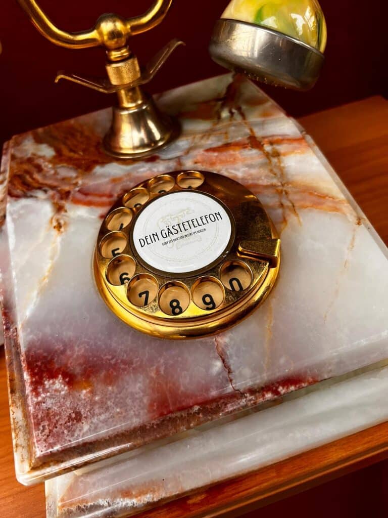 Ein Vintage-Wählscheibentelefon mit goldfarbener Wählscheibe auf einem Marmorsockel verleiht Ihrer Hochzeitsdekoration einen Hauch von Nostalgie. Mit dem deutschen Etikett „DEIN GÄSTETELEFON“ auf einer erdig getönten Steinoberfläche ergänzt es die Eleganz Ihrer Hochzeitsdekoration auf wunderbare Weise.