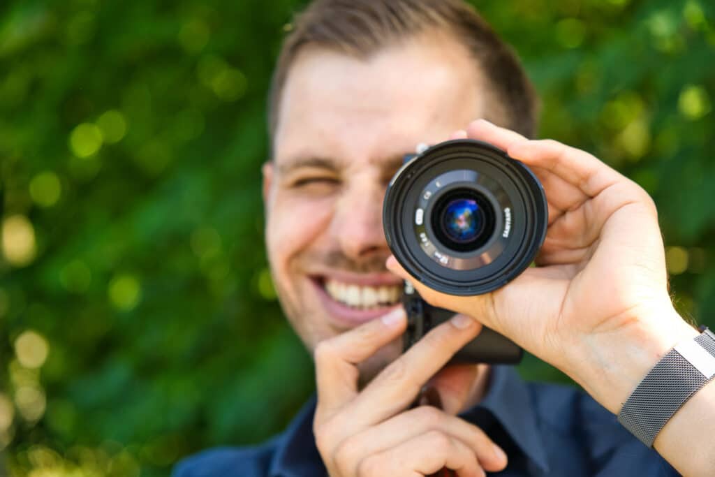 Ein lächelnder Mann hält sich ein Kameraobjektiv vors Auge und imitiert damit ein Monokel inmitten der Hochzeitsvorbereitungen vor einem verschwommenen grünen Hintergrund. Der Fokus liegt auf dem Objektiv, wodurch eine spielerische und kreative Szene entsteht, die die freudige Essenz der Hochzeitsplanung einfängt.