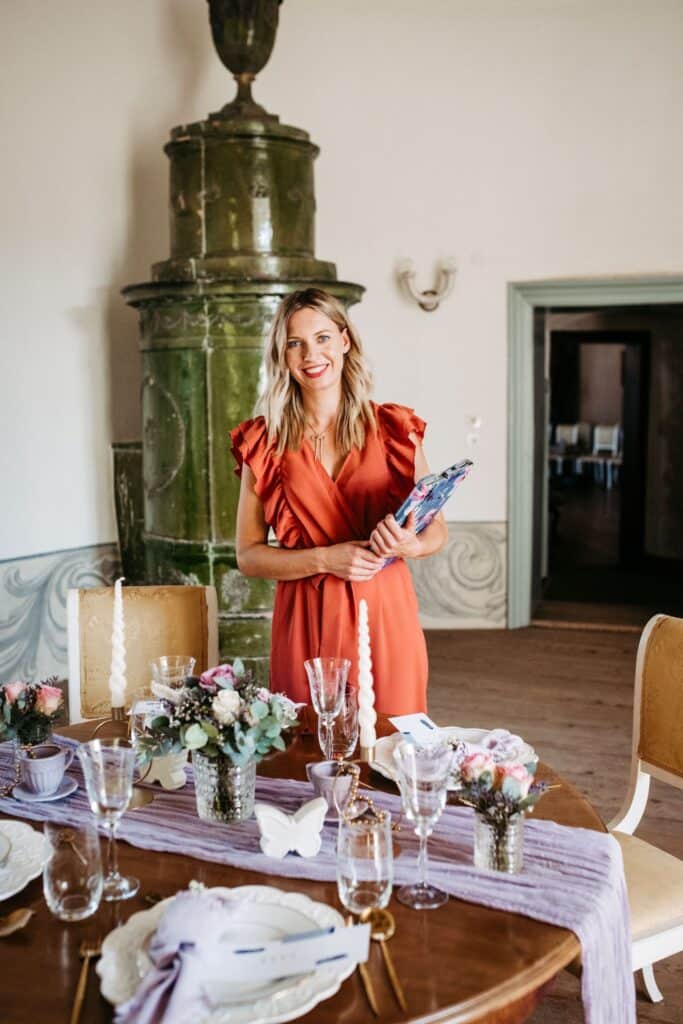 Eine Person in einem roten Kleid steht an einem Esstisch mit Glaswaren, Kerzen und Blumendekorationen in einem elegant dekorierten Raum, der perfekt für eine Hochzeit ist. Im Hintergrund ist ein verzierter grüner Ofen zu sehen. Sie hält eine gefaltete Serviette und lächelt in die Kamera, um die Freude der Hochzeitsplanung einzufangen.