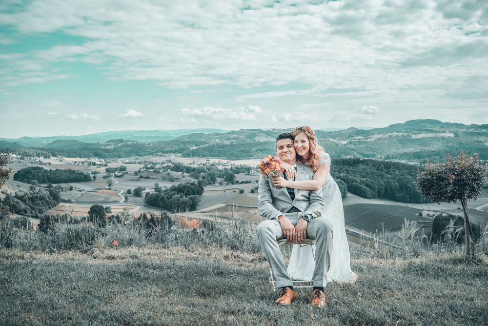 Ein Paar im Hochzeitskleid posiert im Freien vor einer malerischen Landschaft und verkörpert die Schönheit des Heiratens. Der Bräutigam sitzt auf einem Stuhl, während die Braut mit einem Blumenstrauß hinter ihm steht. Sanfte Hügel und Felder erstrecken sich unter einem teilweise bewölkten Himmel.