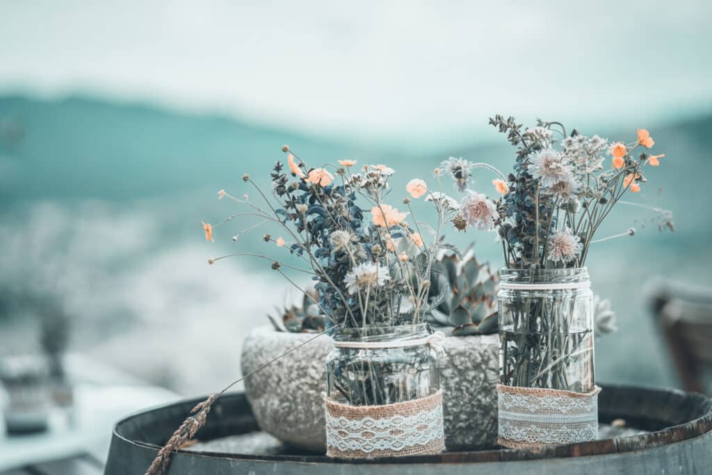 Zwei Töpfe mit zarten Wildblumen, in Spitze eingewickelt, ruhen auf einem Holzfass und rufen eine intime Hochzeit hervor. Im Hintergrund steht ein Steinpflanzgefäß mit Sukkulenten. Die Szene spielt im Freien, eingerahmt von verschwommenen grünen Hügeln unter einem bewölkten Himmel – perfekt für eine zauberhafte Hochzeitskulisse im Freien.