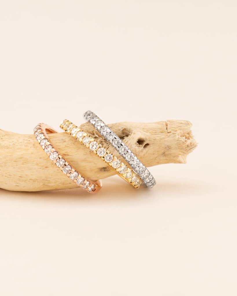 Vier funkelnde Armreifen aus Roségold, Gelbgold und Silber, mit klaren Edelsteinen verziert, werden auf einem beigefarbenen Treibholzstück vor einem hellen Hintergrund präsentiert. Perfekt für alle, die ihre Hochzeit planen, lassen sich die Armbänder elegant stapeln, um ihre schimmernden Steine hervorzuheben.