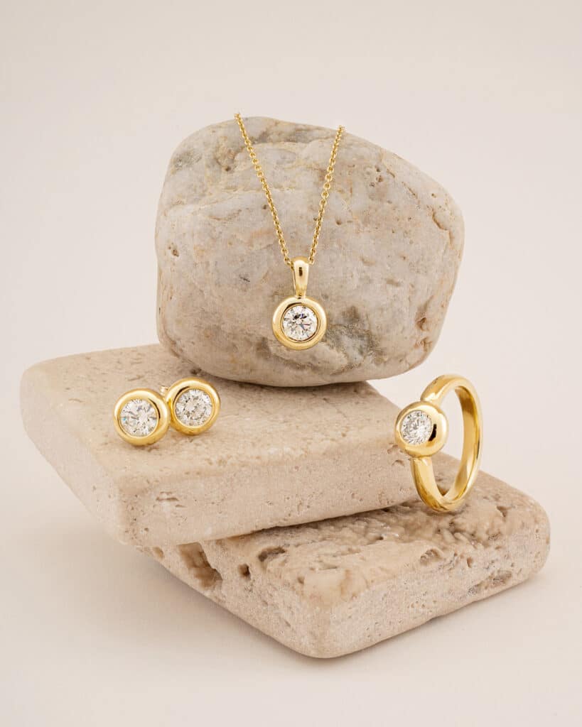Ein Goldschmuckset, perfekt für eine Hochzeit, bestehend aus einer Halskette, einem Ring und Ohrringen, verziert mit runden Diamantakzenten. Diese exquisiten Stücke werden elegant auf glatten beigen Steinen vor einem neutralen Hintergrund präsentiert.