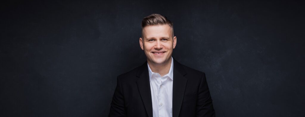 Ein Mann in Anzug und weißem Hemd lächelt vor einem dunklen, strukturierten Hintergrund und strahlt die Freude und Eleganz einer Person aus, die in die Hochzeitsplanung vertieft ist.