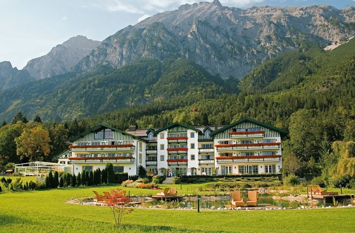 Ein großes Hotel mit farbenfrohen Balkonen liegt in einer üppigen grünen Landschaft, perfekt für Hochzeitsplanung. Im Vordergrund bietet ein kleiner Teich mit Stühlen drumherum einen idealen Ort für intime Momente. Majestätische Berge erheben sich im Hintergrund unter einem klaren blauen Himmel und machen es zu einem Traumort zum Heiraten.
