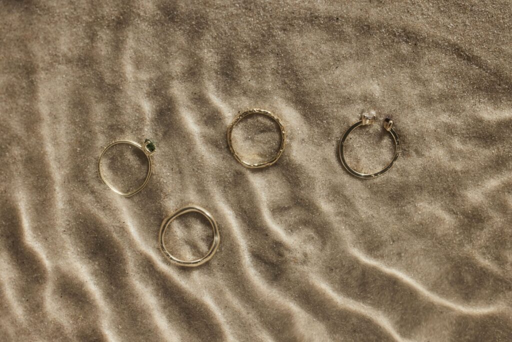 Vier zarte Ringe sind auf einer strukturierten Sandoberfläche angeordnet, perfekt für diejenigen, die ihre Hochzeit planen. Jeder Ring hat ein einzigartiges Design, einer davon mit einem kleinen grünen Edelstein. Das wellenförmige Muster des Sandes wirft subtile Schatten um den Schmuck und spiegelt die Eleganz von Heiraten wider.