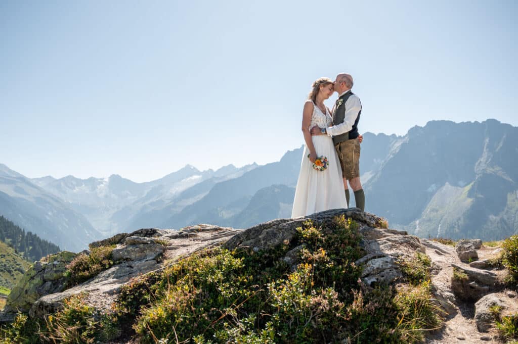 Braut und Bräutigam stehen auf einem felsigen Berggipfel und umarmen sich vor der Kulisse majestätischer Berge unter einem klaren blauen Himmel. Während sie ihre Hochzeit feiern, hält die Braut einen Blumenstrauß in den Händen, der die Schönheit ihrer Verbindung inmitten der Erhabenheit der Natur symbolisiert.