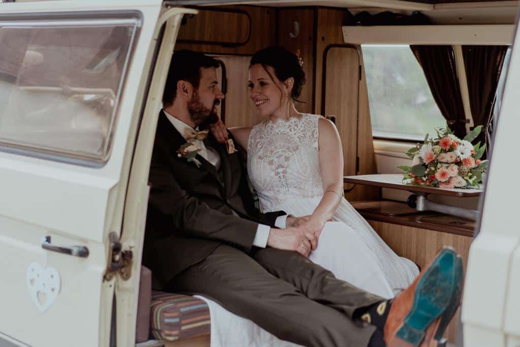 Braut und Bräutigam sitzen in einem Oldtimer-Van, lächeln sich an und verkörpern die Freude einer Hochzeit. Die Braut trägt ein weißes Spitzenkleid und der Bräutigam einen dunklen Anzug. Im Hintergrund steht ein Blumenstrauß auf einem Tisch und spiegelt die Essenz einer durchdachten Hochzeitsplanung wider.