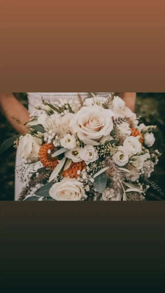 Eine Person hält einen großen, eleganten Blumenstrauß mit cremefarbenen Rosen, weißen Blumen, orangefarbenen Akzenten und grünem Laub – eine perfekte Note für die Hochzeitsplanung – vor einem unscharfen Hintergrund.