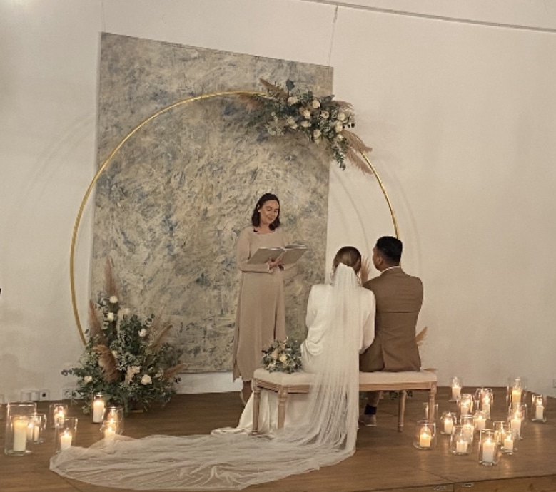 Ein Paar sitzt auf einer Bank gegenüber dem Trauungsbeamten. Sie sind von Kerzen und einem runden Blumenbogen umgeben, der mit Pampasgras geschmückt ist, perfekt für ihre Hochzeit. Die Braut trägt einen langen Schleier und ein weißes Kleid, während der Bräutigam einen hellbraunen Anzug trägt.