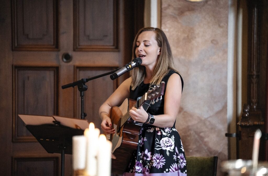 In einem warm beleuchteten Raum singt eine Frau in einem geblümten Kleid und spielt Akustikgitarre in ein Mikrofon. Kerzen verschwimmen im Vordergrund und schaffen eine zauberhafte Kulisse, die sich perfekt für romantische Momente wie die Hochzeitsplanung eignet.