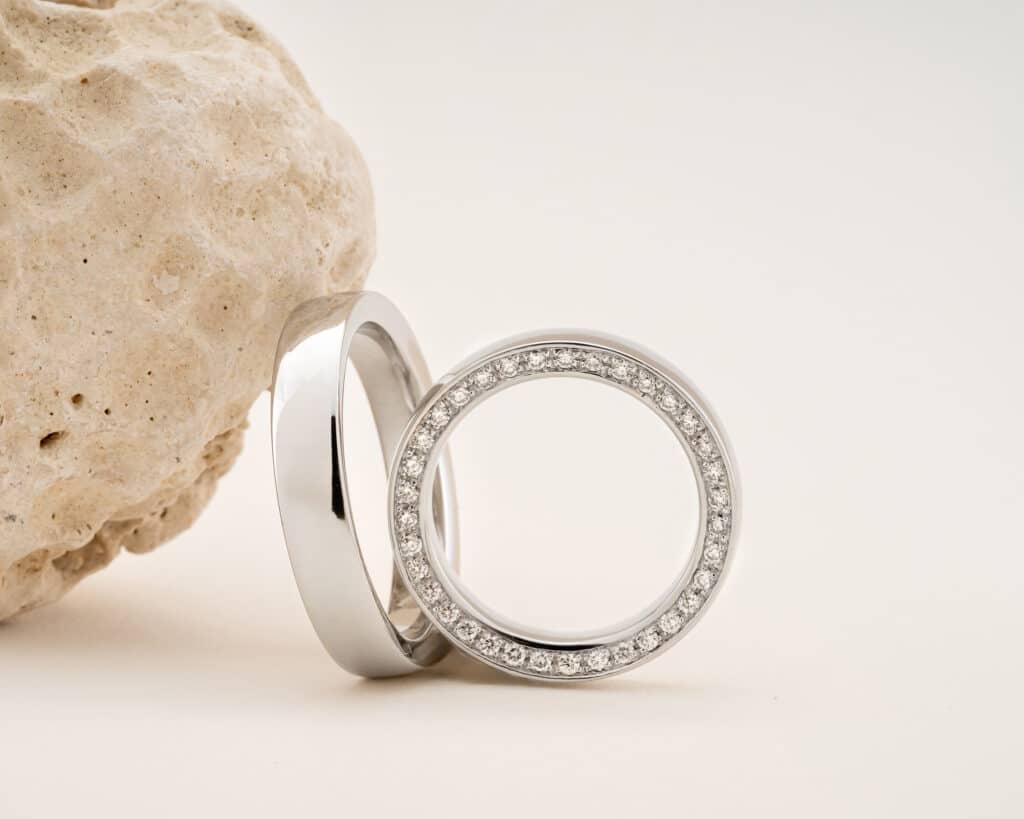Zwei Silberringe, perfekt für eine Hochzeit, liegen neben einem rauen, strukturierten hellen Stein. Ein Ring hat ein schlichtes Band, während der andere ein kreisförmiges Design aufweist, das durch kleine Diamanten am Rand hervorgehoben wird – ideale Symbole für diejenigen, die heiraten möchten.