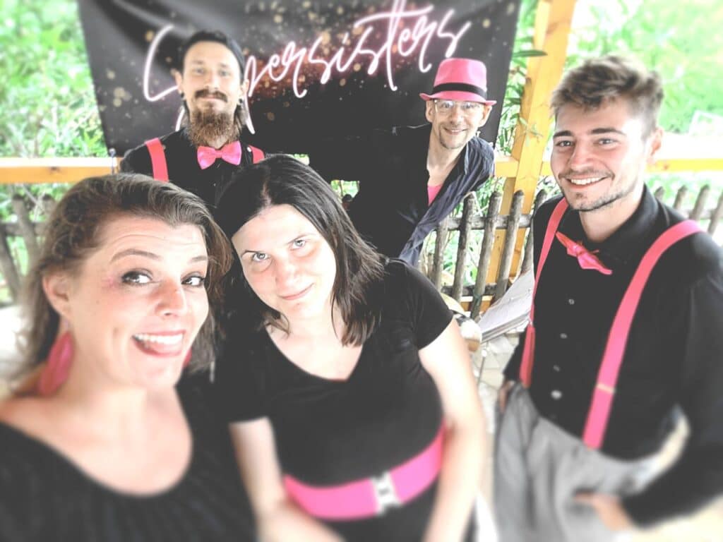 Fünf lächelnde Menschen vor einem „Coversisters“-Banner, gekleidet in schwarze Outfits mit leuchtend rosa Akzenten wie Hüten, Hosenträgern und Fliegen. Die perfekte Inspiration für alle, die an der Hochzeitsplanung beteiligt sind, mit Grünpflanzen und einem Holzzaun, der einen Hauch von rustikalem Charme verleiht.