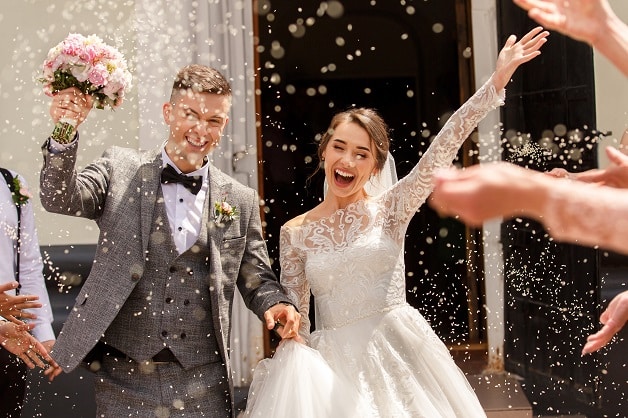 Ein fröhliches Brautpaar feiert seine Hochzeit vor einem Gebäude. Der Bräutigam hält einen Blumenstrauß und beide lächeln breit. Die Gäste überschütten sie mit Konfetti, während sie Hand in Hand gehen und die Braut aufgeregt winkt.