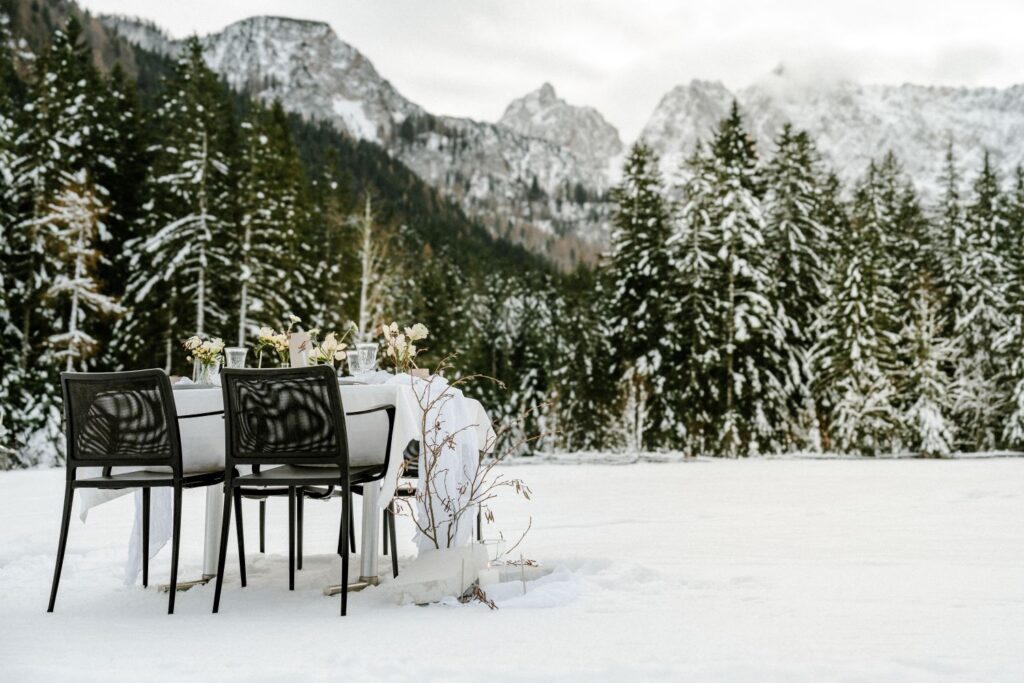 Ein für ein Essen gedeckter Tisch steht im Freien in einer verschneiten Landschaft, perfekt für eine intime Hochzeit. Umgeben von schneebedeckten Bäumen und Bergen wertet die Szene die Hochzeitsplanung mit weißen und gelben Blumen auf einer weißen Tischdecke auf, ergänzt durch elegante schwarze Stühle.