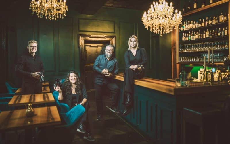 Eine Gruppe von vier Personen sitzt in einer schwach beleuchteten Bar mit dunkelgrünen Wänden und posiert lässig um eine hölzerne Bartheke. Zwei Kronleuchter hängen von der Decke und verbreiten ein romantisches Licht – perfekt, um Hochzeitspläne zu besprechen. Regale voller Flaschen tragen zur Atmosphäre bei, während sie ihre Hochzeit feiern wollen.