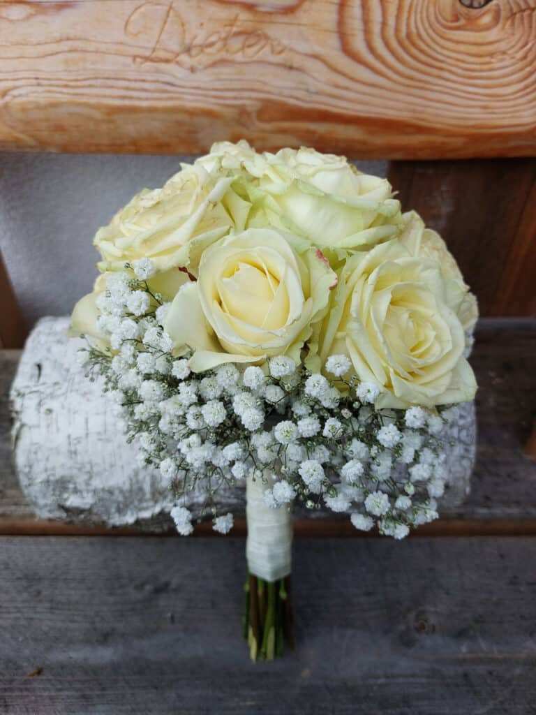Ein Strauß mit blassgelben Rosen, umgeben von zartem weißem Schleierkraut, umwickelt mit einer weißen Schleife, perfekt für Ihre Hochzeitsplanung. Das Arrangement ruht wunderschön auf einer rustikalen Holzoberfläche.