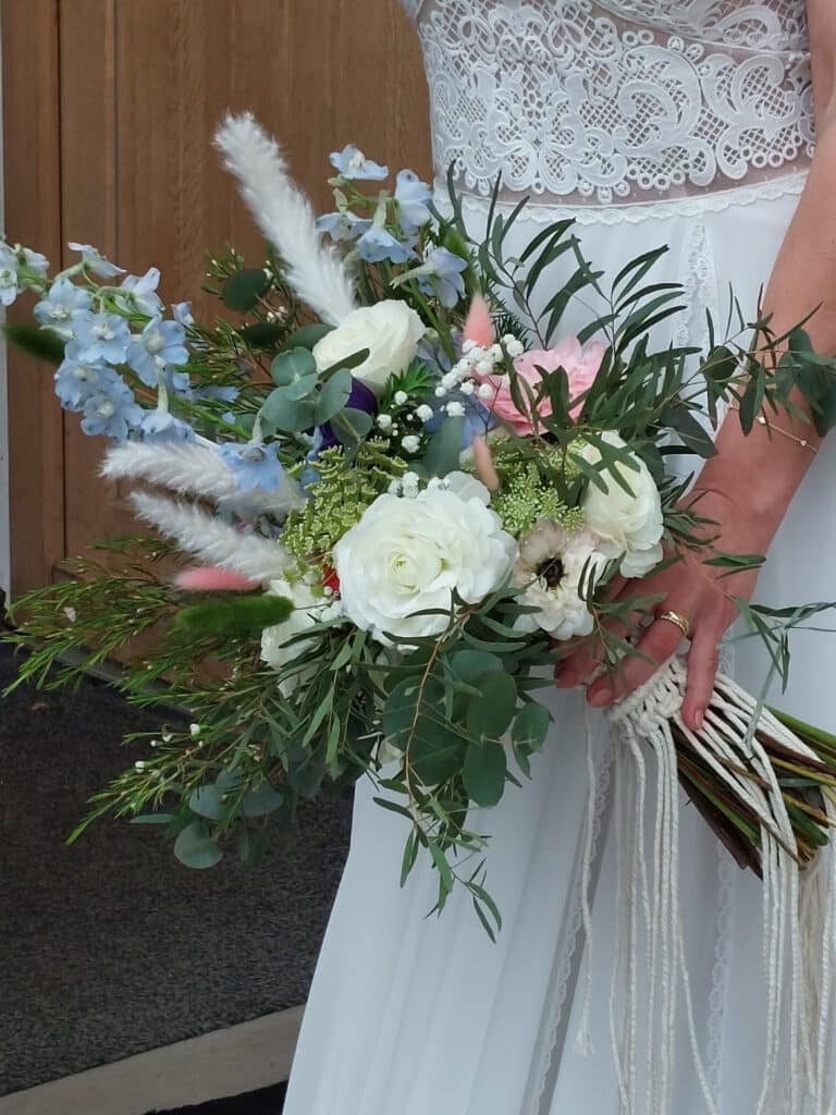 Eine Braut hält einen großen, farbenfrohen Blumenstrauß mit weißen Rosen, blauen Ritterspornen und rosa Akzenten inmitten üppigen Grüns. Sie trägt ein elegantes weißes Spitzenkleid vor dem bezaubernden Hintergrund einer Holztür – und fängt damit wahrhaftig die Essenz einer Traumhochzeit ein.