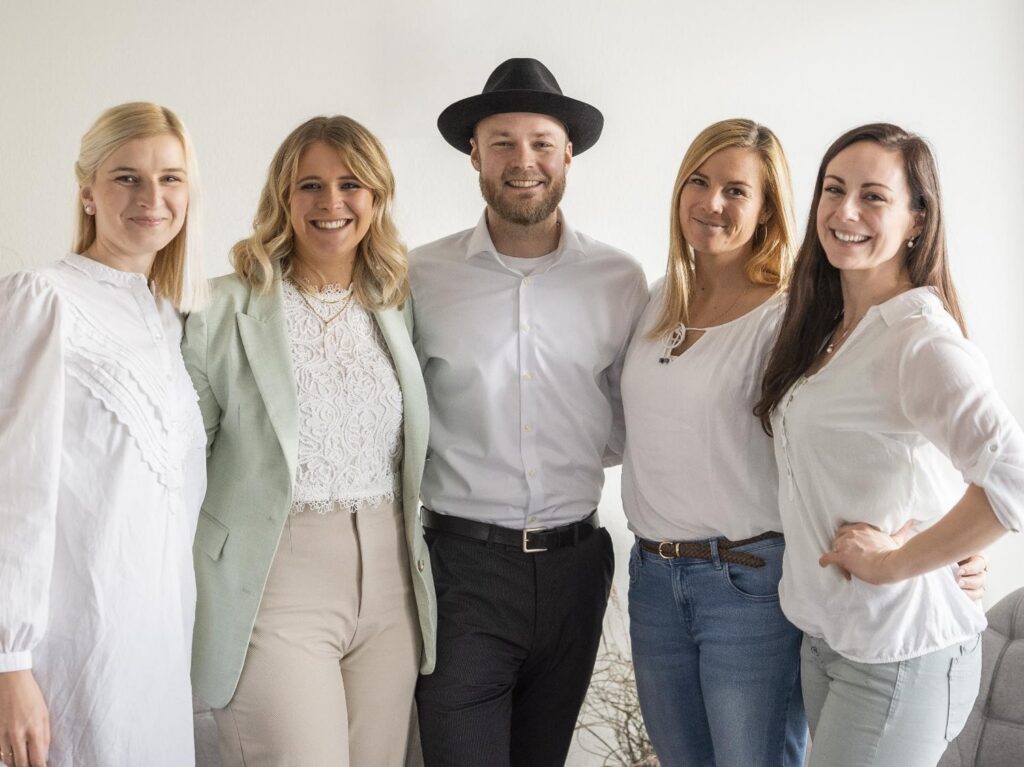Eine Gruppe von fünf Personen steht zusammen im Haus und strahlt in die Kamera. Drei Frauen links, ein Mann mit Hut in der Mitte und eine Frau rechts. Alle sind in heller Freizeitkleidung gekleidet und bereit, sich in die Hochzeitsplanung zu stürzen.