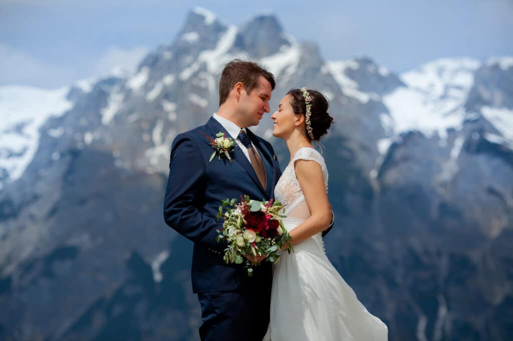 Braut und Bräutigam stehen eng beieinander und schauen sich an, im Hintergrund schneebedeckte Berge. Die Braut hält einen Strauß weißer und roter Blumen in der Hand, der ihre wunderschöne Hochzeit symbolisiert. Beide tragen formelle Hochzeitskleidung, die Monate sorgfältiger Hochzeitsplanung verkörpert.