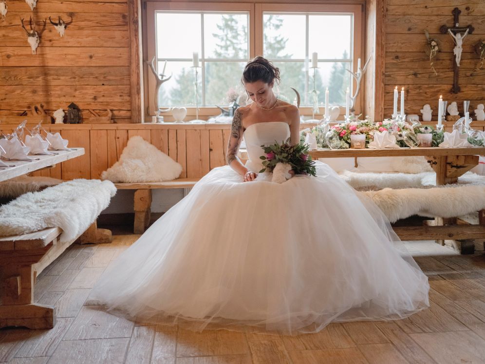 Eine Braut in einem weißen Kleid sitzt auf einer rustikalen Holzbank in einer gemütlichen Hütte und denkt über ihre Hochzeitsplanung nach. Sie hält einen Blumenstrauß in der Hand und blickt nach unten, umgeben von flauschigen Pelzdecken und Blumendekorationen, während ein großes Fenster ruhiges Licht ins Innere bringt.