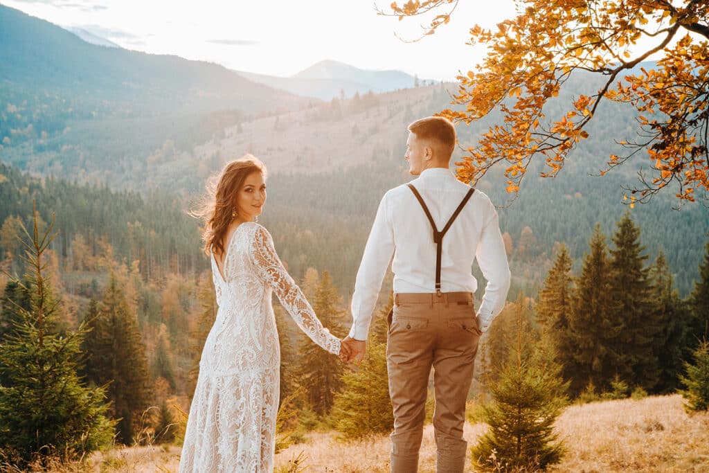 Ein Paar steht mit verschränkten Händen auf einem Hügel und verkörpert damit die Essenz von Heiraten. Die Frau trägt ein weißes Spitzenkleid und der Mann ein weißes Hemd mit Hosenträgern. Sie blicken einander vor einer Kulisse aus malerischen Bergen und leuchtendem Herbstlaub an.