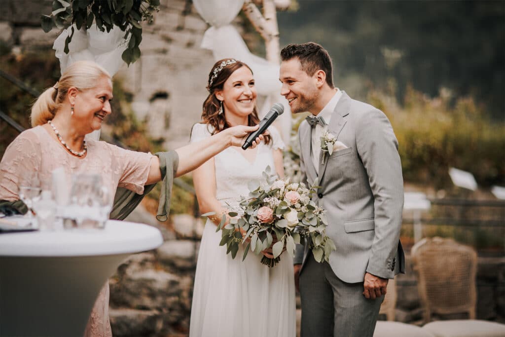 Während ihrer Hochzeit im Freien strahlen Braut und Bräutigam vor Freude. Der Bräutigam spricht in ein Mikrofon, das eine Frau hält. Die Braut in ihrem weißen Kleid hält einen Strauß rosa und weißer Blumen. Die natürliche Umgebung mit üppigem Grün rundet diesen wunderschönen Moment ab.