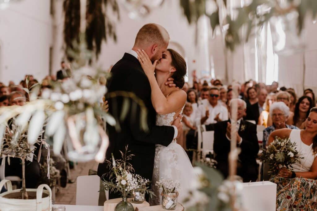 Ein frisch vermähltes Paar küsst sich in einem wunderschön dekorierten Hochzeitssaal, umgeben von Gästen. Die Braut trägt ein atemberaubendes weißes Kleid und der Bräutigam ist elegant gekleidet. Die Gäste sitzen in Reihen und feiern den Moment, während farbenfrohe Blumenarrangements die romantische Atmosphäre verstärken.