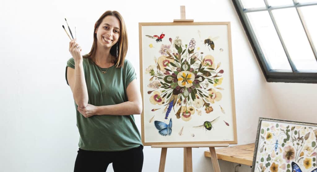 Eine Frau in einem grünen Hemd steht lächelnd neben einer Staffelei und präsentiert ihre Kunstwerke mit Blumen- und Insektenmotiven – perfekt für alle, die nach einzigartigen Dekorationsideen für eine Hochzeit suchen. Sie hält drei Pinsel in einer Hand, im Hintergrund ist teilweise ein Fenster sichtbar.
