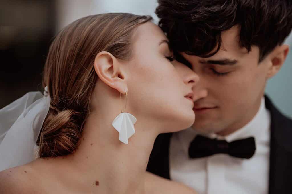 Braut und Bräutigam, eingefangen in einem intimen Moment während ihrer Hochzeit, tauschen einen zärtlichen Blick. Die einzigartigen Ohrringe der Braut funkeln unter ihrem weißen Schleier, während sie sich zu dem Bräutigam in seinem schwarzen Smoking beugt. Vor einem sanft verschwommenen Hintergrund wirken sie nah und liebevoll.