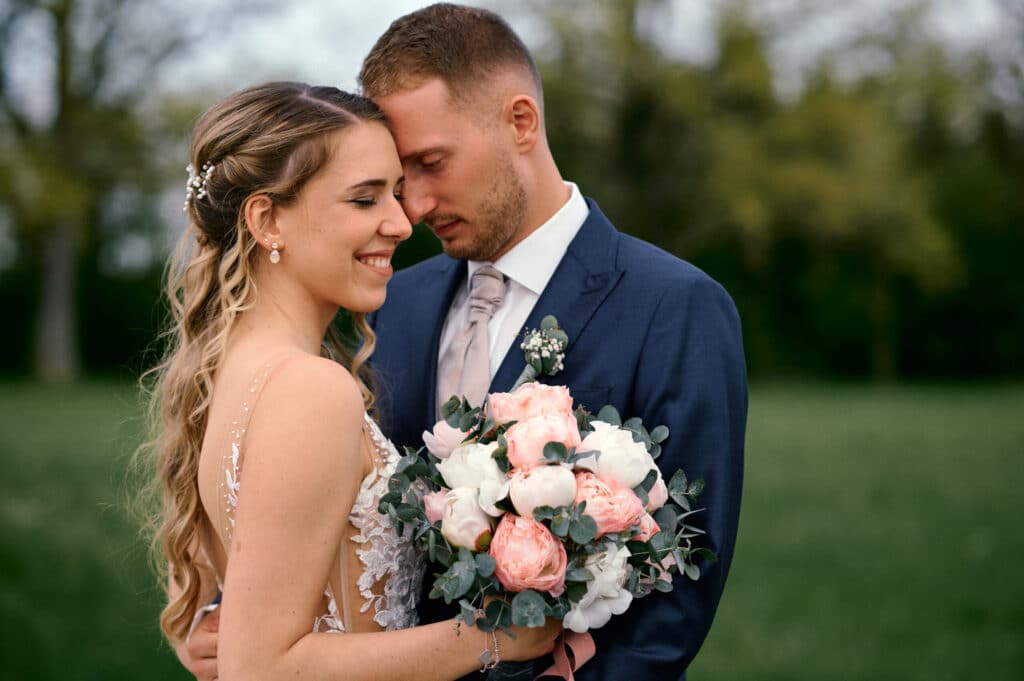 Braut und Bräutigam stehen eng beieinander im Freien, vereint in ihrer Hochzeit. Die Braut, lächelnd, mit langem, welligem Haar, hält einen Strauß weißer und rosa Blumen. Der Bräutigam, in einem blauen Anzug, lehnt seinen Kopf sanft an ihren und hat geschlossene Augen. Im Hintergrund sind Bäume verschwommen.