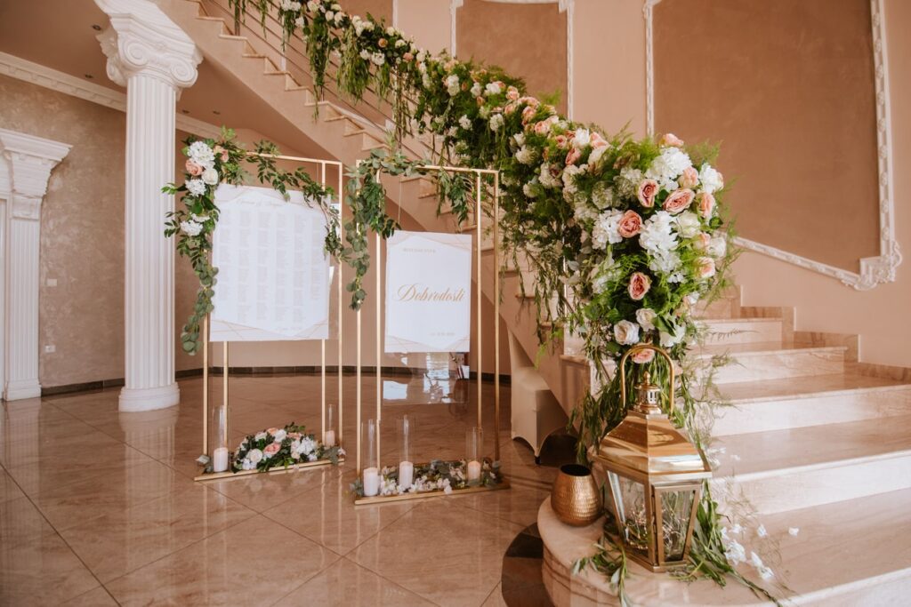 Ein dekorierter Hochzeitseingang mit eleganten Blumenarrangements auf der Treppe, bestehend aus weißen und rosa Blumen und Grünpflanzen. Zwei goldene Ständer mit Sitzplänen sind ausgestellt und eine große goldene Laterne steht auf dem Marmorboden und schafft eine perfekte Kulisse für die Hochzeit.