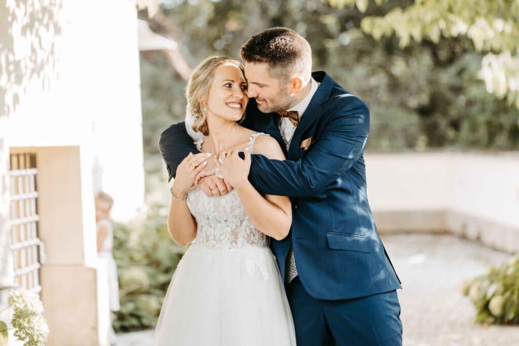 Ein Paar im Hochzeitskleid genießt einen freudigen Moment im Freien. Die Braut in einem weißen Spitzenkleid und der Bräutigam in einem blauen Anzug umarmen sich unter einem Blätterdach. Sie lächeln sich in der sonnigen Umgebung an und leben ihren Hochzeitstraum.