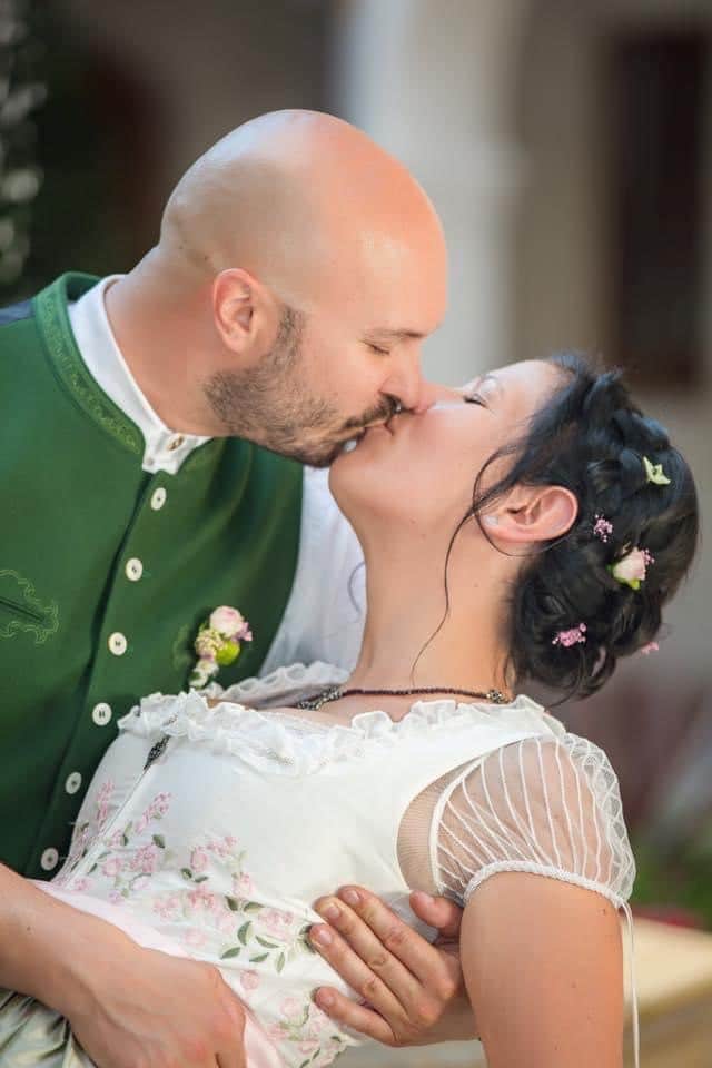 Ein Paar küsst sich in traditioneller Kleidung und fängt einen romantischen Hochzeitsmoment ein. Der Mann trägt eine grüne Weste mit Blumendetails und die Frau ein weißes Kleid mit rosa Blumen und transparenten Ärmeln. In ihr geflochtenes Haar sind Blumen eingeflochten, während sie sich leicht nach hinten lehnt.