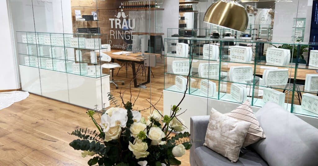 Ein gemütliches, modernes Juweliergeschäft mit Holzböden und weißen Wänden strahlt eine Atmosphäre von Eleganz aus. Glasvitrinen präsentieren Ringe, die perfekt für die Hochzeitsgäste sind. Ein Strauß weißer Rosen ziert den Vordergrund, während ein Sofa mit Kissen und eine metallische Hängelampe die Szenerie vervollständigen.