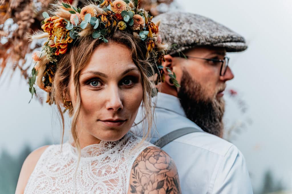 Nahaufnahme eines Paares, das Rücken an Rücken steht und die Glückseligkeit des Heiratens ausstrahlt. Die Frau trägt einen Blumenkranz und ein Spitzenkleid mit einem Blumentattoo auf der Schulter, während der Mann eine Brille, eine Mütze und ein weißes Hemd trägt. Der Hintergrund ist weich und unscharf.