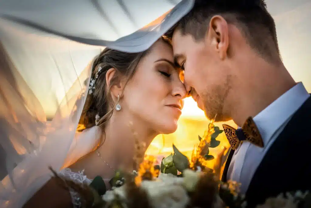 Braut und Bräutigam berühren sanft ihre Stirnen unter einem durchsichtigen Schleier und genießen die Wärme des Sonnenuntergangs an ihrem Hochzeitstag. Umgeben von Liebe und Blumen fängt diese intime Szene die Romantik des Heiratens perfekt ein.