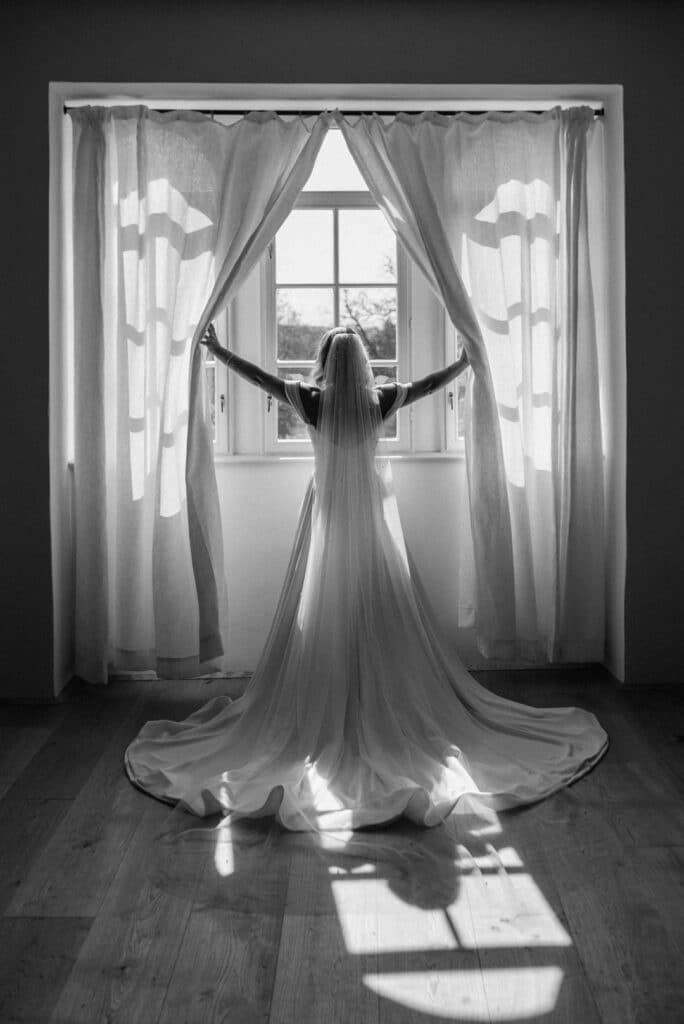 Eine Braut steht vor einem Fenster und hält die Vorhänge offen. Sie trägt ein langes Brautkleid mit Schleppe und Schleier und verkörpert damit ihren perfekten Hochzeitstraum. Licht strömt herein und erzeugt eine dramatische Silhouette auf dem Holzboden – ein magischer Moment in der Reise der Hochzeitsplanung.